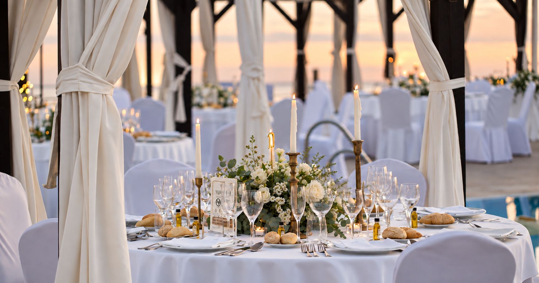 Elegant event table with candles and white flowers set under draped curtains at sunset by the sea